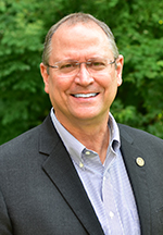 A smiling middle-aged man in a suit stands in front of a lush, green background of trees and foliage